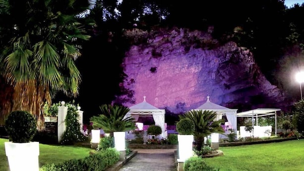 Un jardín con un sendero que lleva a un pabellón, rodeado de abundante vegetación y una formación rocosa iluminada en morado.