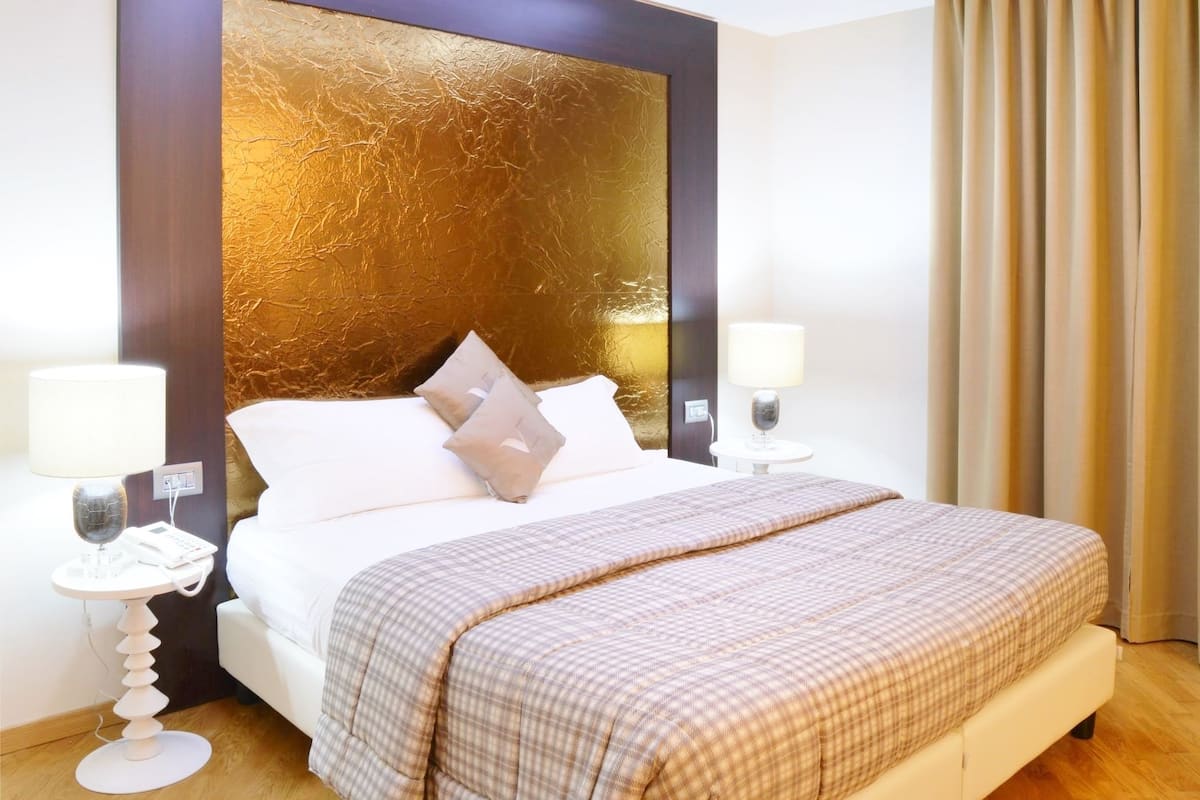 Quarto de hotel com uma cama grande, duas mesas de cabeceira com abajures, uma cabeceira moderna e vista para o exterior através de cortinas.