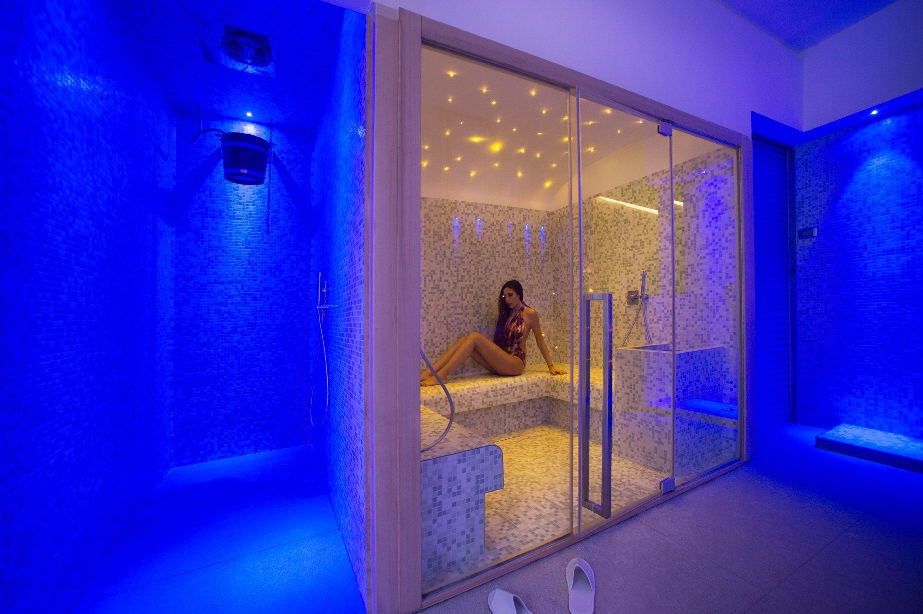 A spa room with a glass partition, a tiled floor, and a person sitting on a bench.
