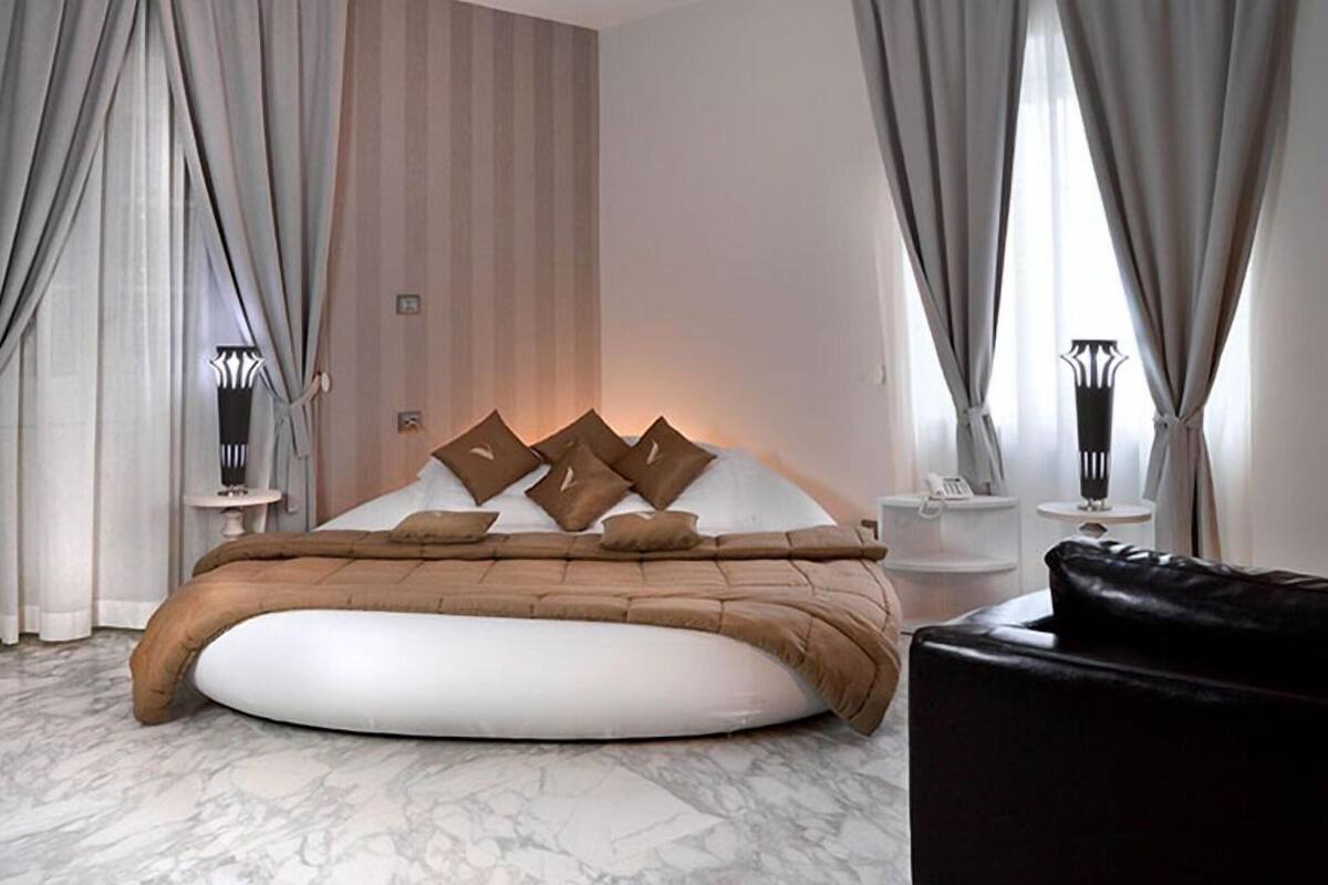 Quarto com uma cama grande, um banquinho redondo e cortinas.