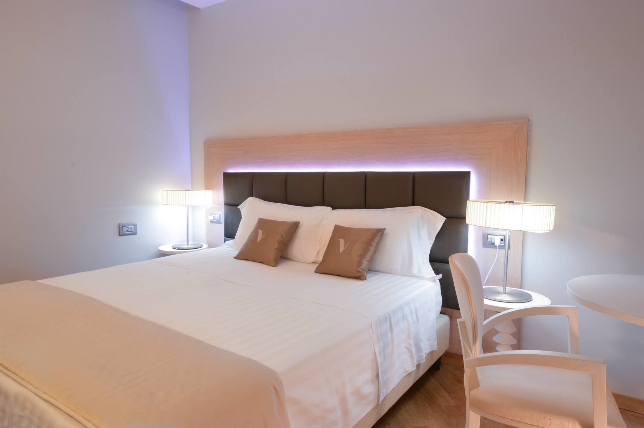 Una cama bien hecha con sábanas blancas y dos almohadas marrones, una mesita de noche con una lámpara, y una pequeña mesa redonda con una silla.
