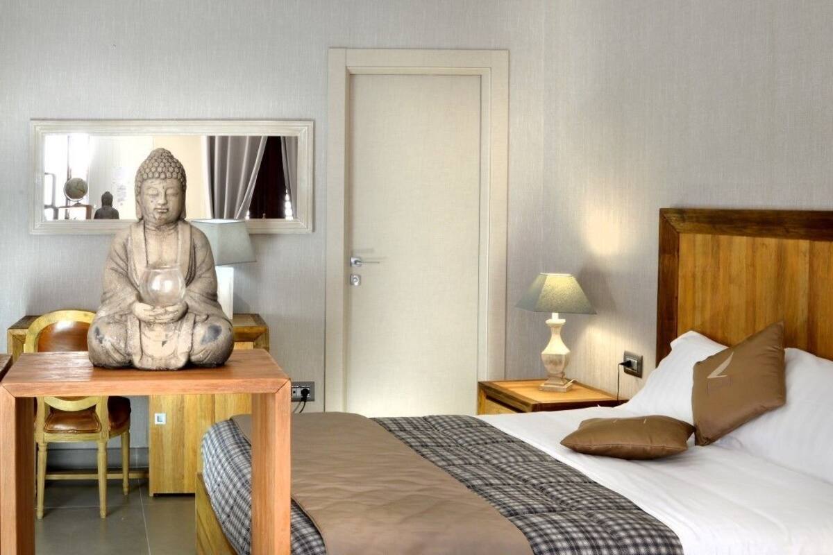 Quarto de hotel com cama, cabeceira de madeira, mesa de cabeceira com luminária, escrivaninha com cadeira, estátua de Buda e espelho na parede.