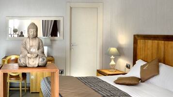 Habitación de hotel con cama, cabecera de madera, mesita de noche con lámpara, escritorio con silla, estatua de Buda y espejo en la pared.