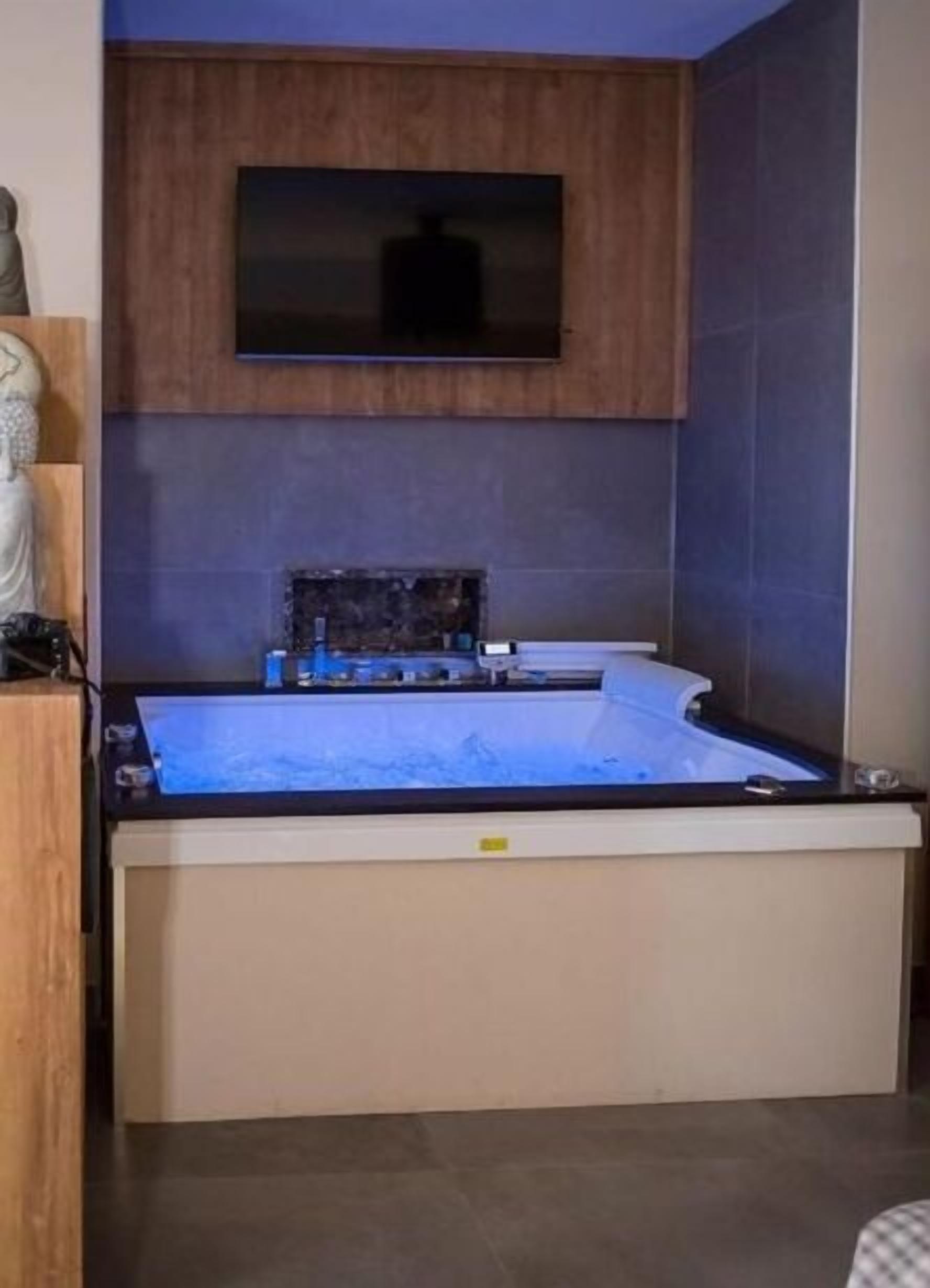 A modern bathroom with a large bathtub, a wooden cabinet, and a flat-screen TV mounted above the tub.