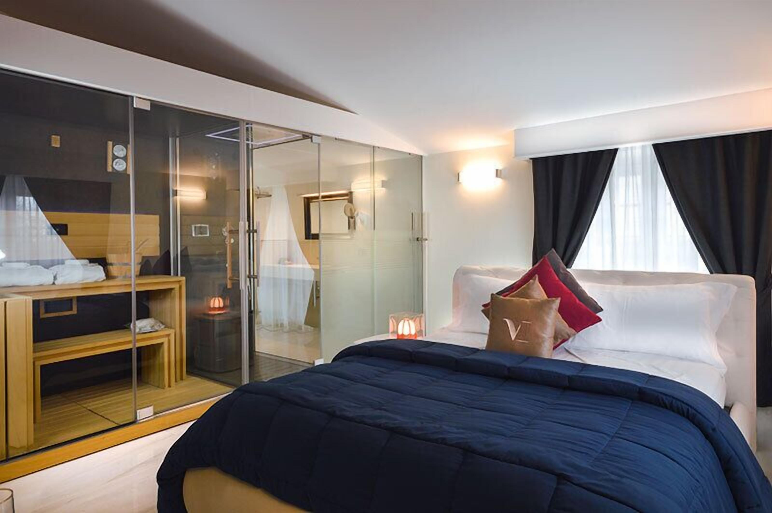A modern hotel room with a glass-enclosed shower, a bed with a blue comforter, and a wooden bench with towels.