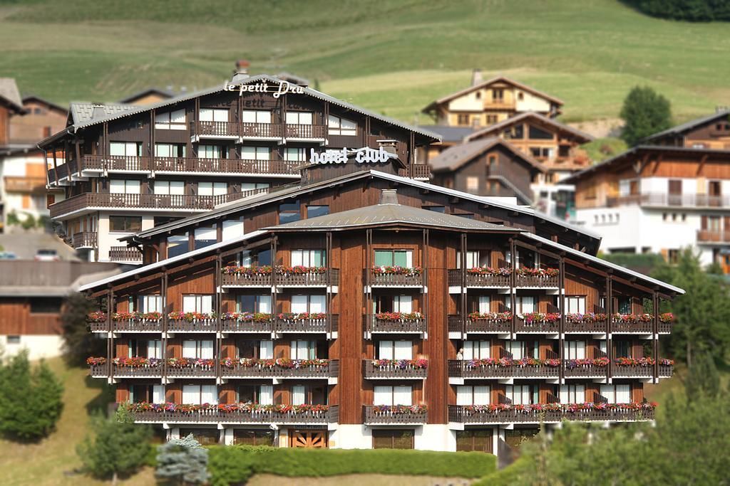 Un hôtel à plusieurs étages doté de balcons, une enseigne indiquant « Hôtel Club », et un paysage montagneux en arrière-plan.