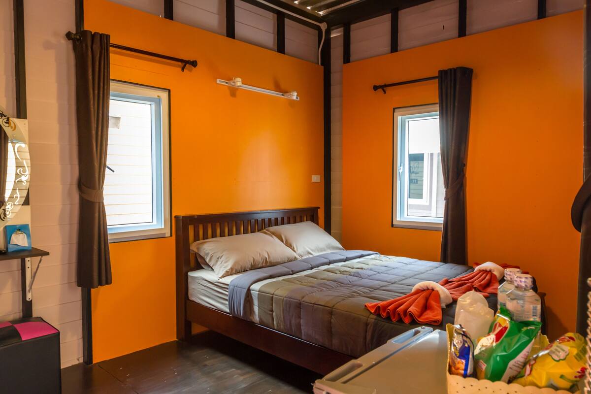 Ein Zimmer mit oranger Wand, einem hölzernen Bett, zwei mit Vorhängen versehenen Fenstern, einem Regal mit einer Tasche und einem Koffer auf dem Boden.
