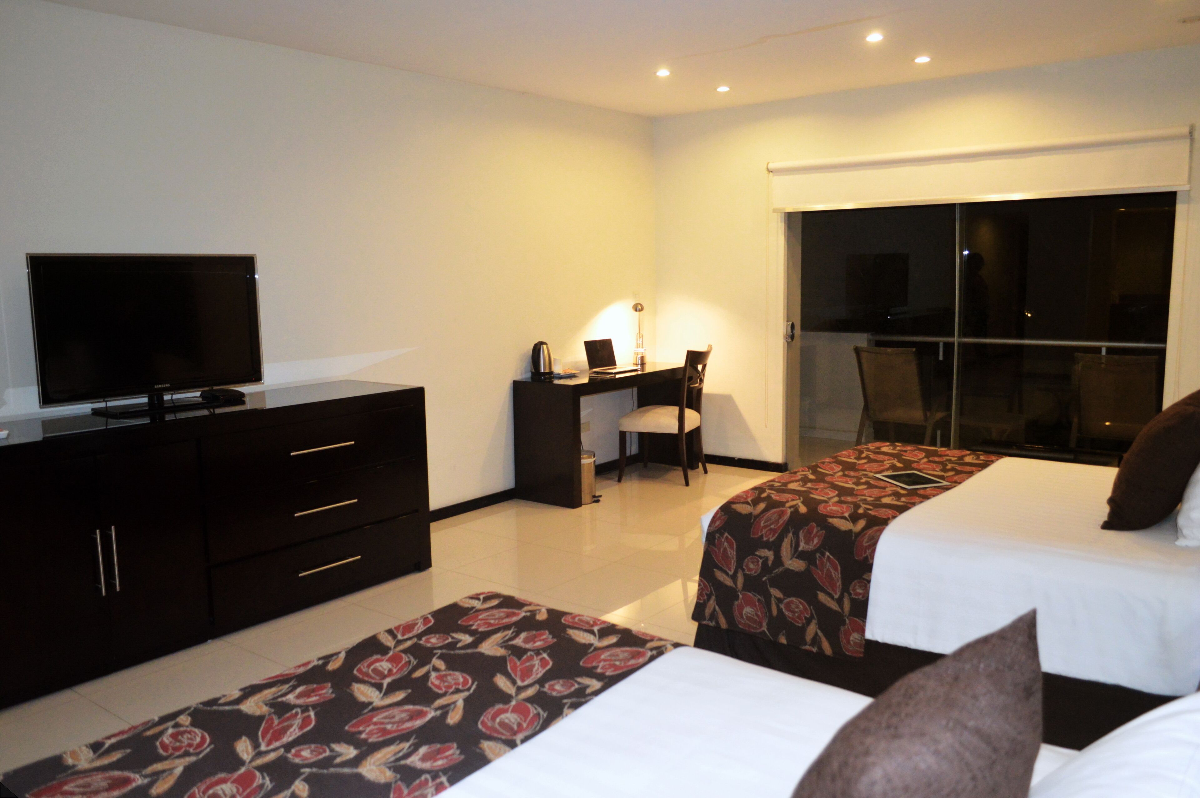 A hotel room with two beds, a desk with a laptop, a television, and a sliding door to a balcony.