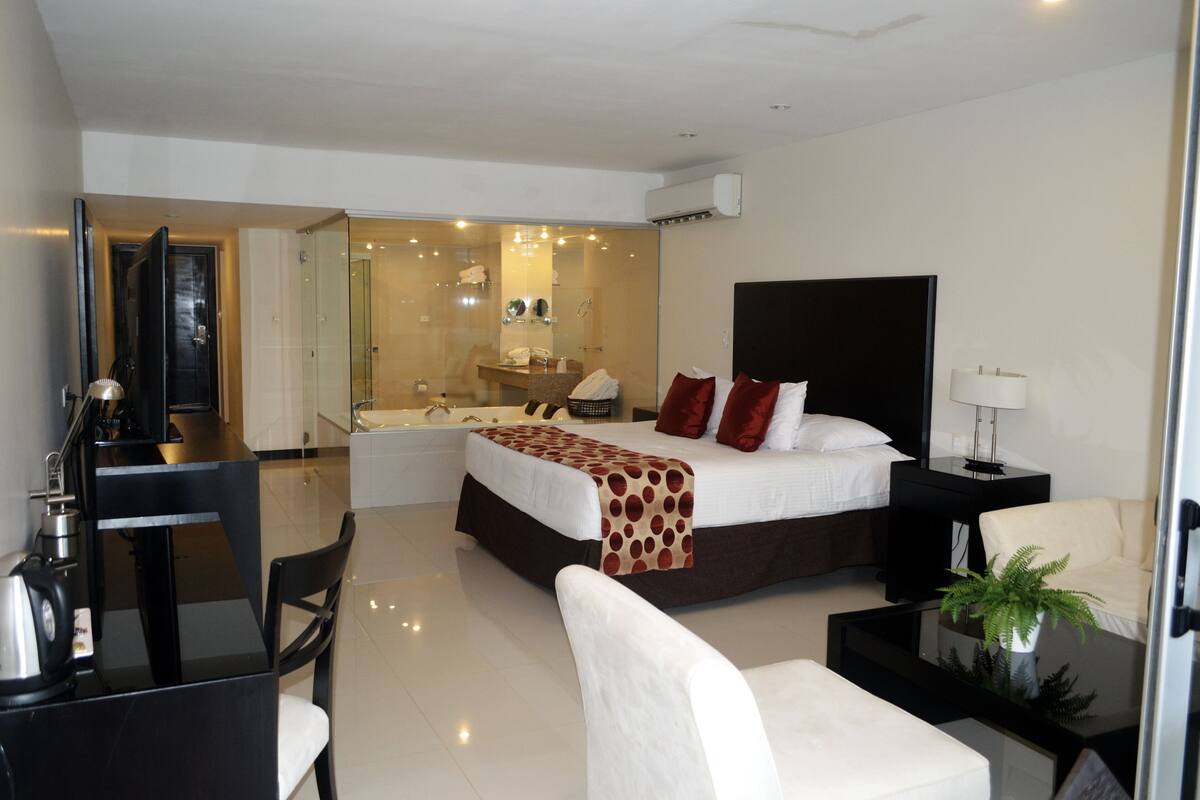 A modern hotel room with a large bed, a desk, a chair, a bedside table with a lamp, and a potted plant.