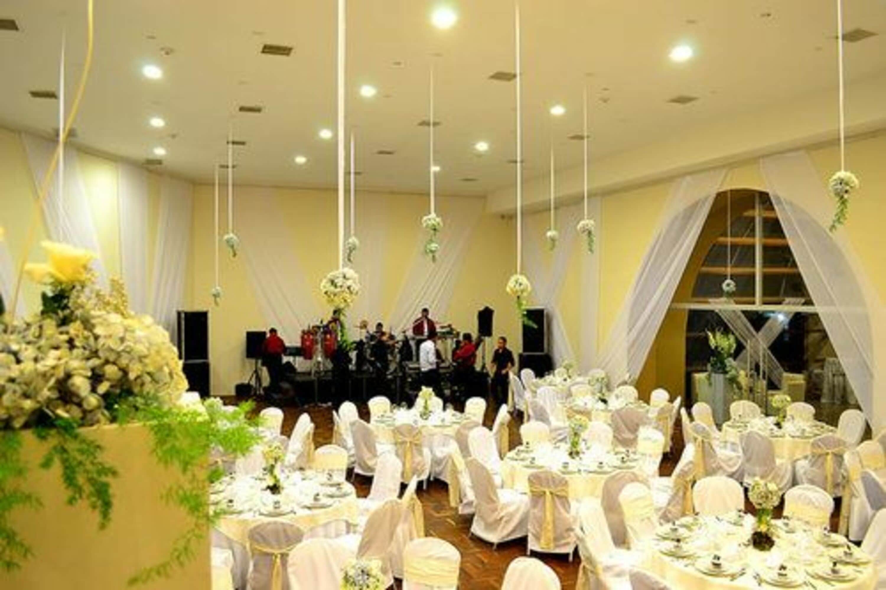 A banquet hall with round tables set for a formal event, draped with white tablecloths and chairs, and decorated with floral arrangements.