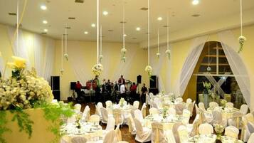 Una sala da banchetti con tavoli rotondi apparecchiati per un evento formale, ricoperti da tovaglie bianche e con sedie, decorata con composizioni floreali.
