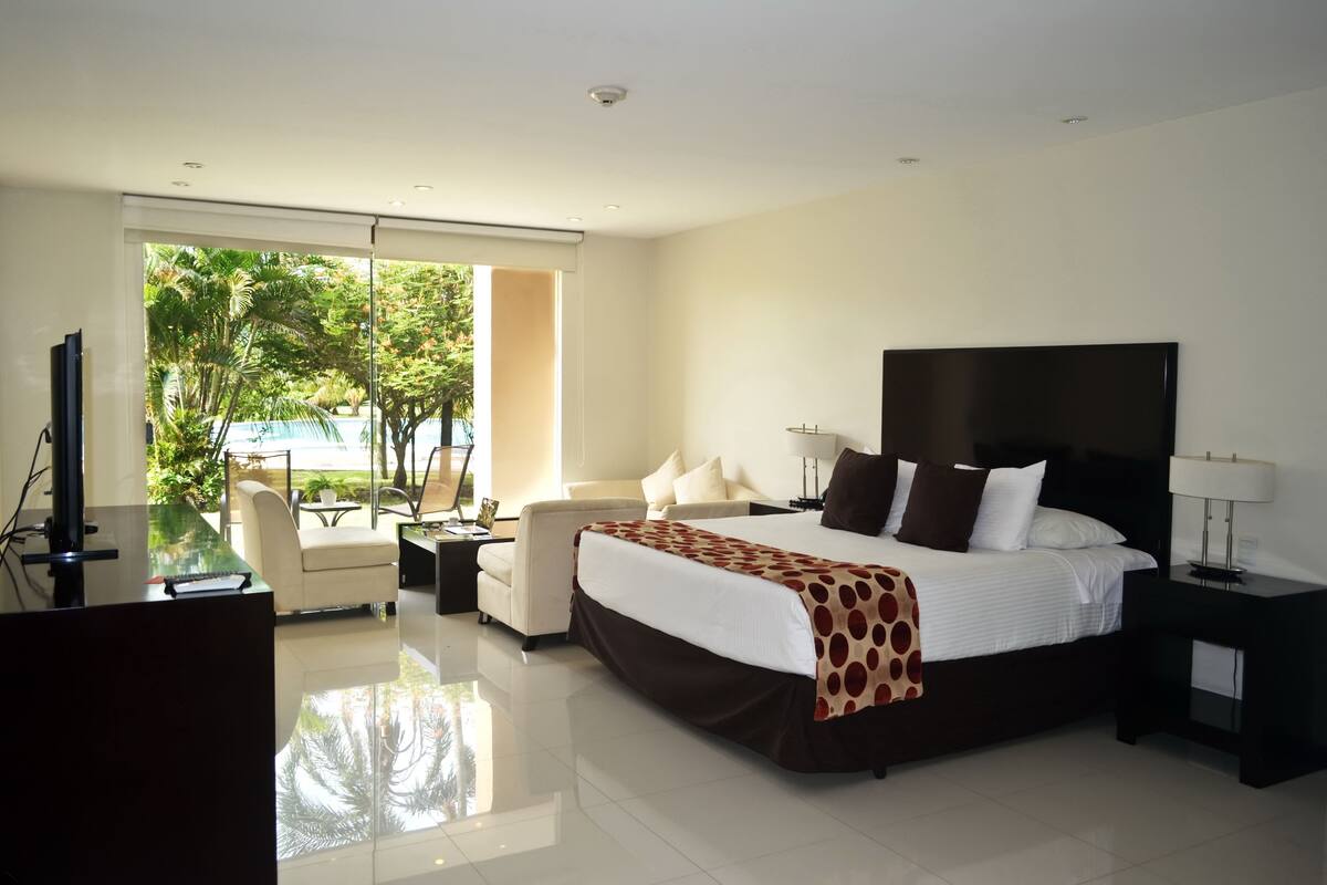 A modern hotel room with a large bed, a desk, a chair, and a view of greenery outside.