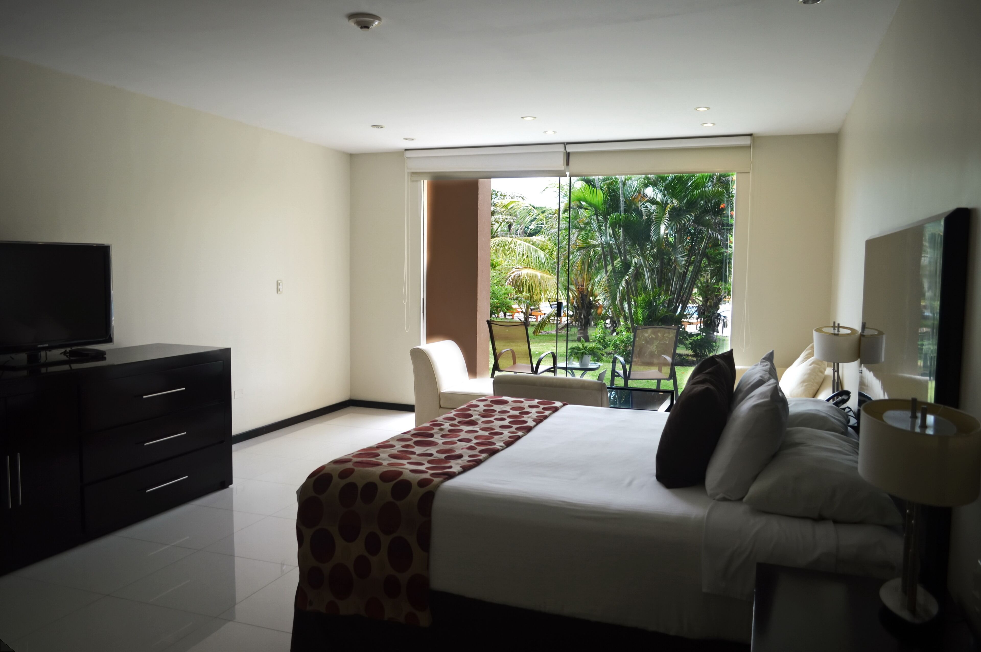 A hotel room with a bed, a TV, a desk, and a window with a view of greenery.