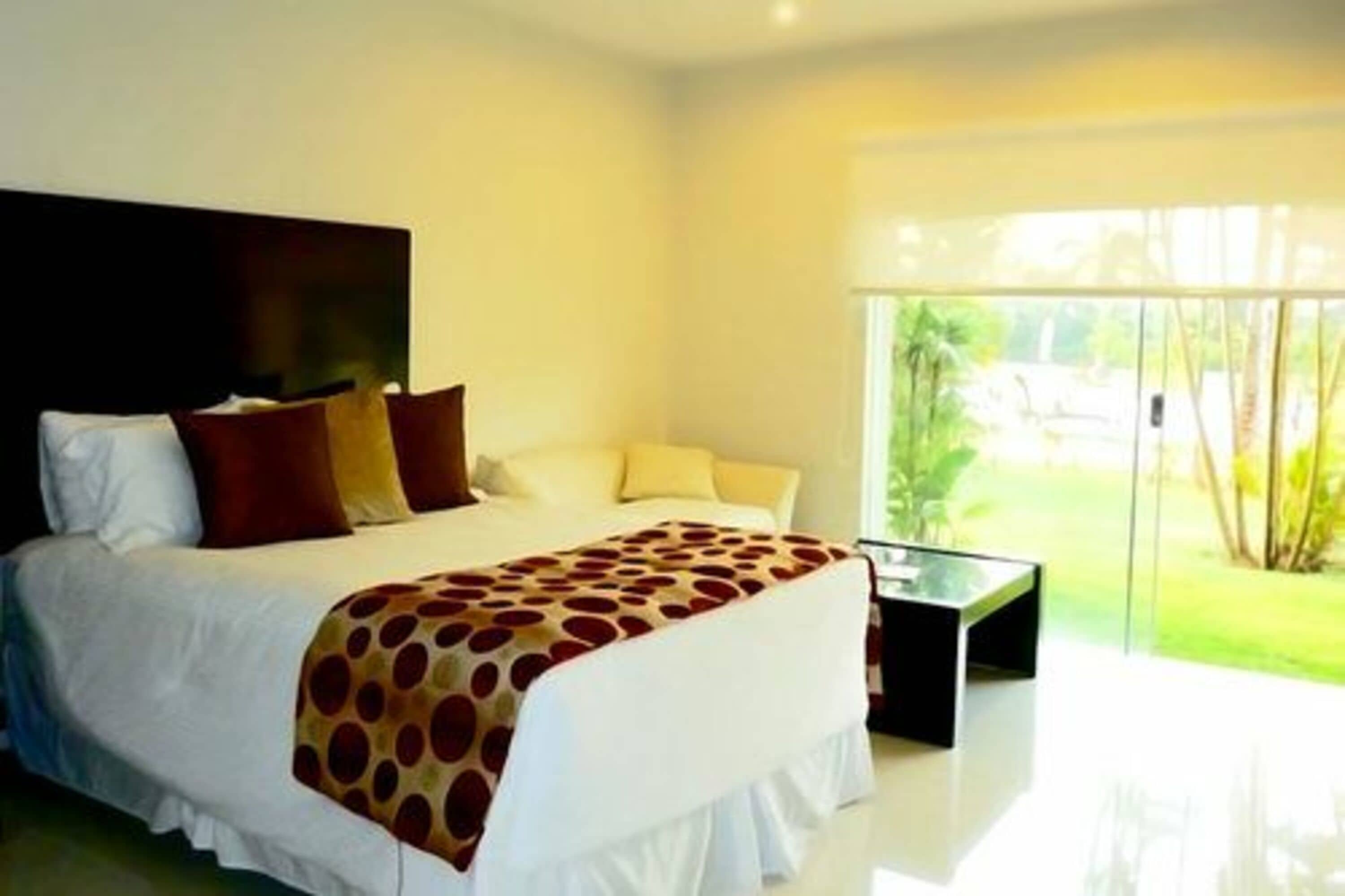 A modern bedroom with a large bed, a glass coffee table, and a view of a garden through large windows.