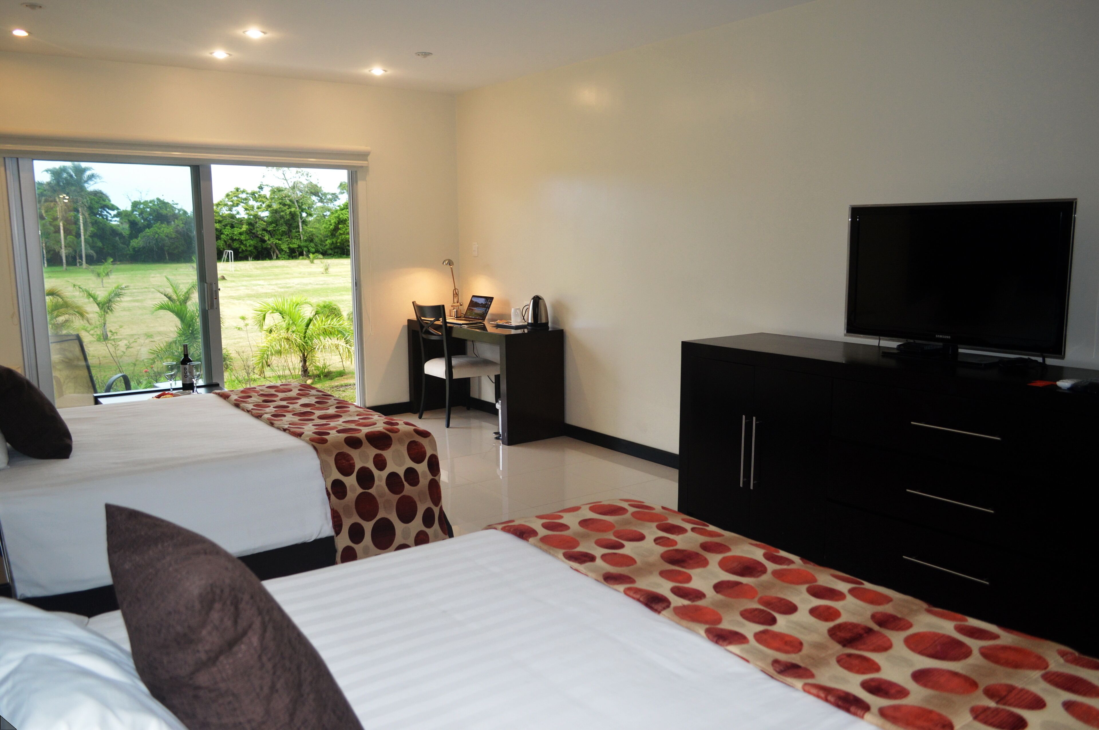 Une chambre d’hôtel avec deux lits, un bureau avec un ordinateur portable, une télévision et une vue sur un jardin, accessible par une porte coulissante.