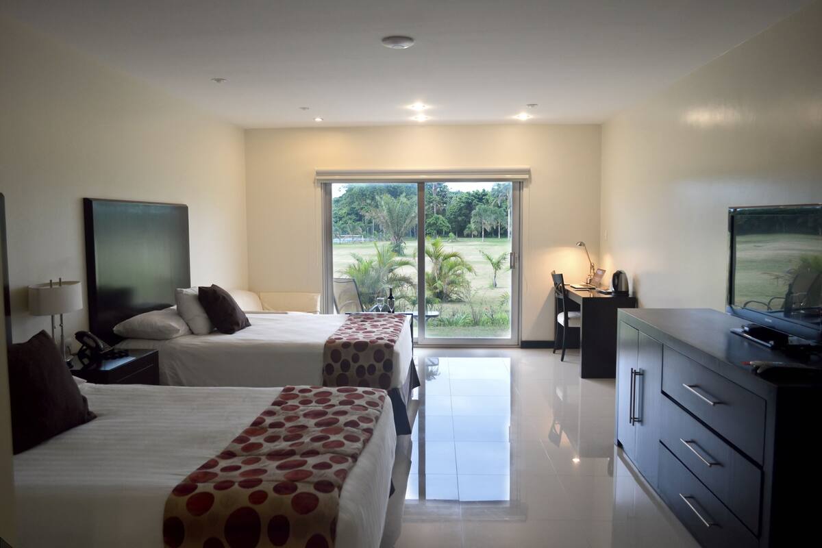 A hotel room with two beds, a desk, a television, and a view of a garden through a sliding door.