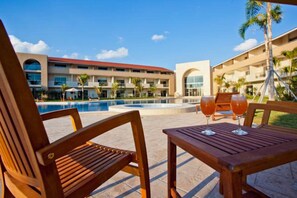 A poolside area with wooden chairs and a table, a pool, and a building with multiple balconies. - Sun Hotel (Santa Cruz)