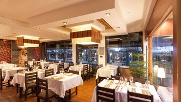 A restaurant with multiple tables set for dining, featuring a modern interior with wooden accents and large windows offering a view of the cityscape.