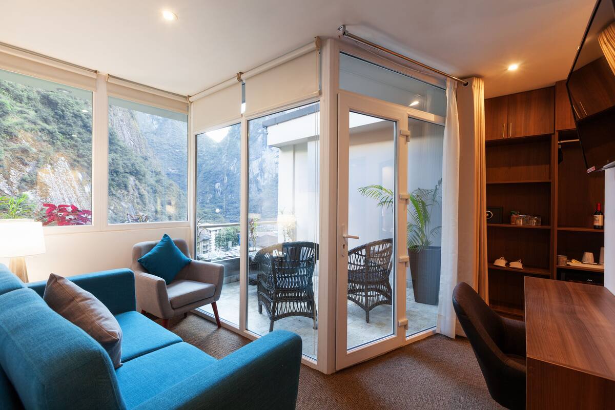 Ein modernes Wohnzimmer mit einer Couch, Sesseln, einem Holztisch und einer Glastür, die auf einen Balkon mit Gartenmöbeln führt.