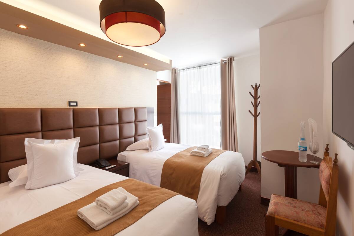 Ein Hotelzimmer mit zwei Betten, einem Holzkopfende, einem kleinen Tisch mit Stuhl und einem großen Fernseher an der Wand.