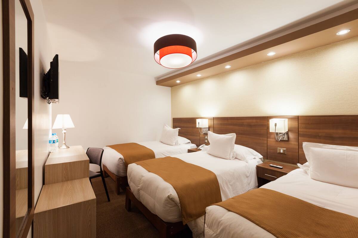 Ein Hotelzimmer mit zwei Betten, einem an der Wand montierten Fernseher, einem Schreibtisch mit Lampe und einem hölzernen Kopfteil.