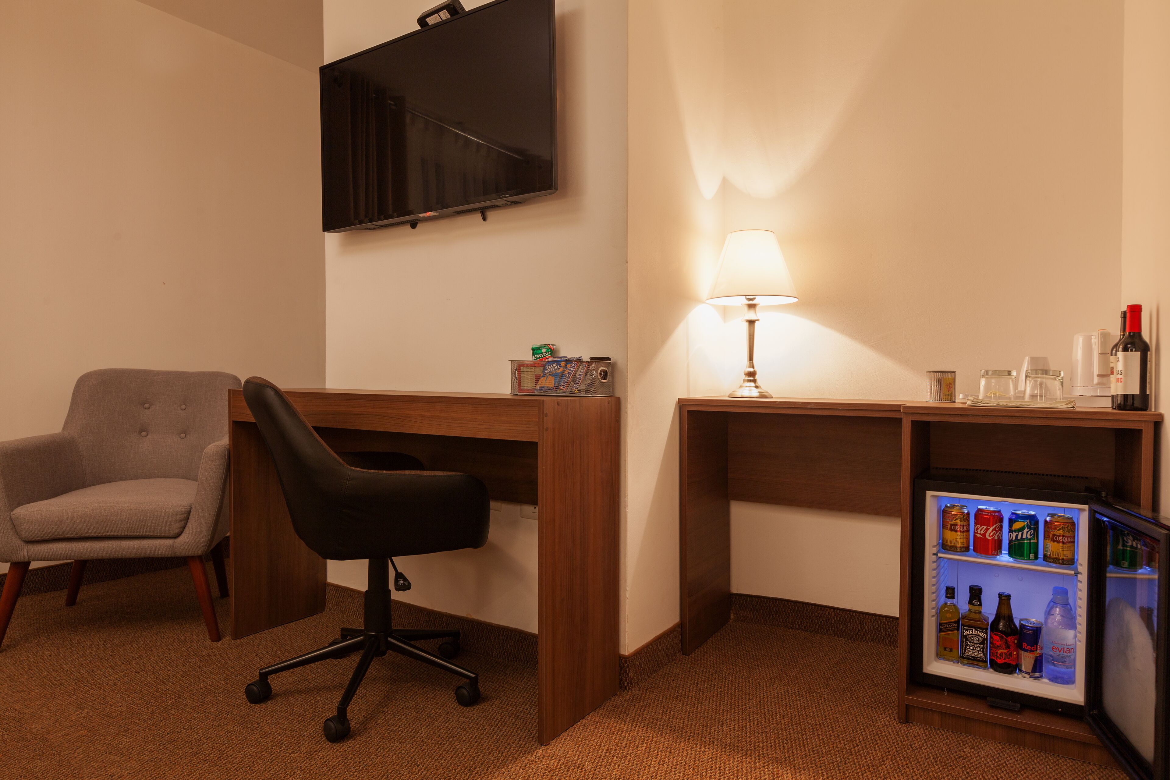 A hotel room with a desk, a chair, a television mounted on the wall, a lamp, and a mini-fridge with beverages.