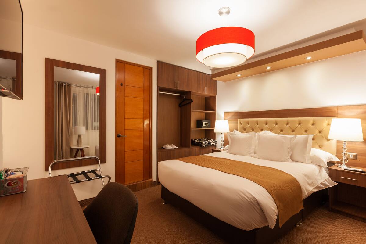 Ein Hotelzimmer mit einem großen Bett, einem Holzschrank, einem Schreibtisch mit Stuhl, einem Spiegel und einer roten Pendelleuchte.