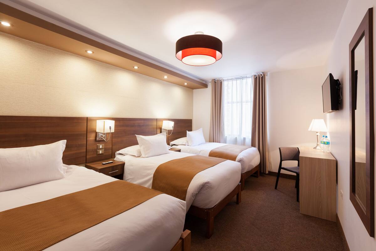 Ein Hotelzimmer mit drei Betten, einem Schreibtisch, einem Stuhl und einem an der Wand befestigten Fernseher.