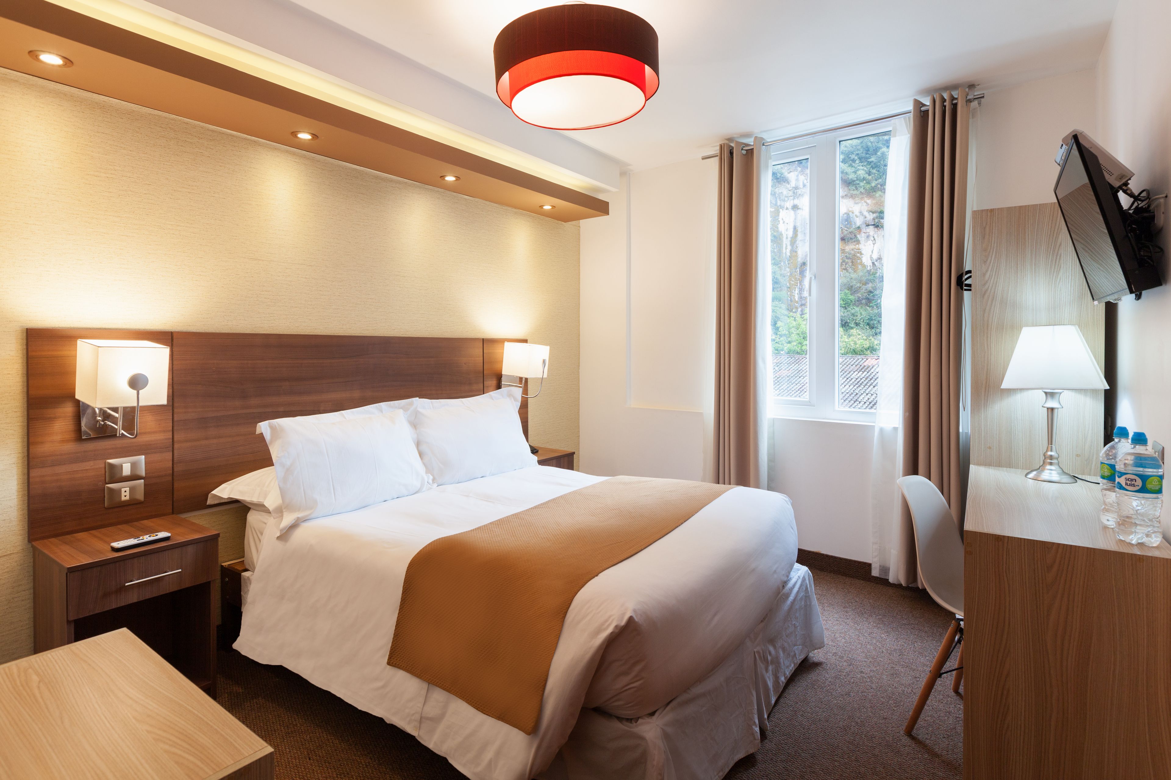 A hotel room with a large bed, bedside lamps, a desk with a TV, and a window with curtains.