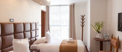 Habitación de hotel con dos camas, cabecera de madera, una mesita con un florero y una botella de agua, y una silla.