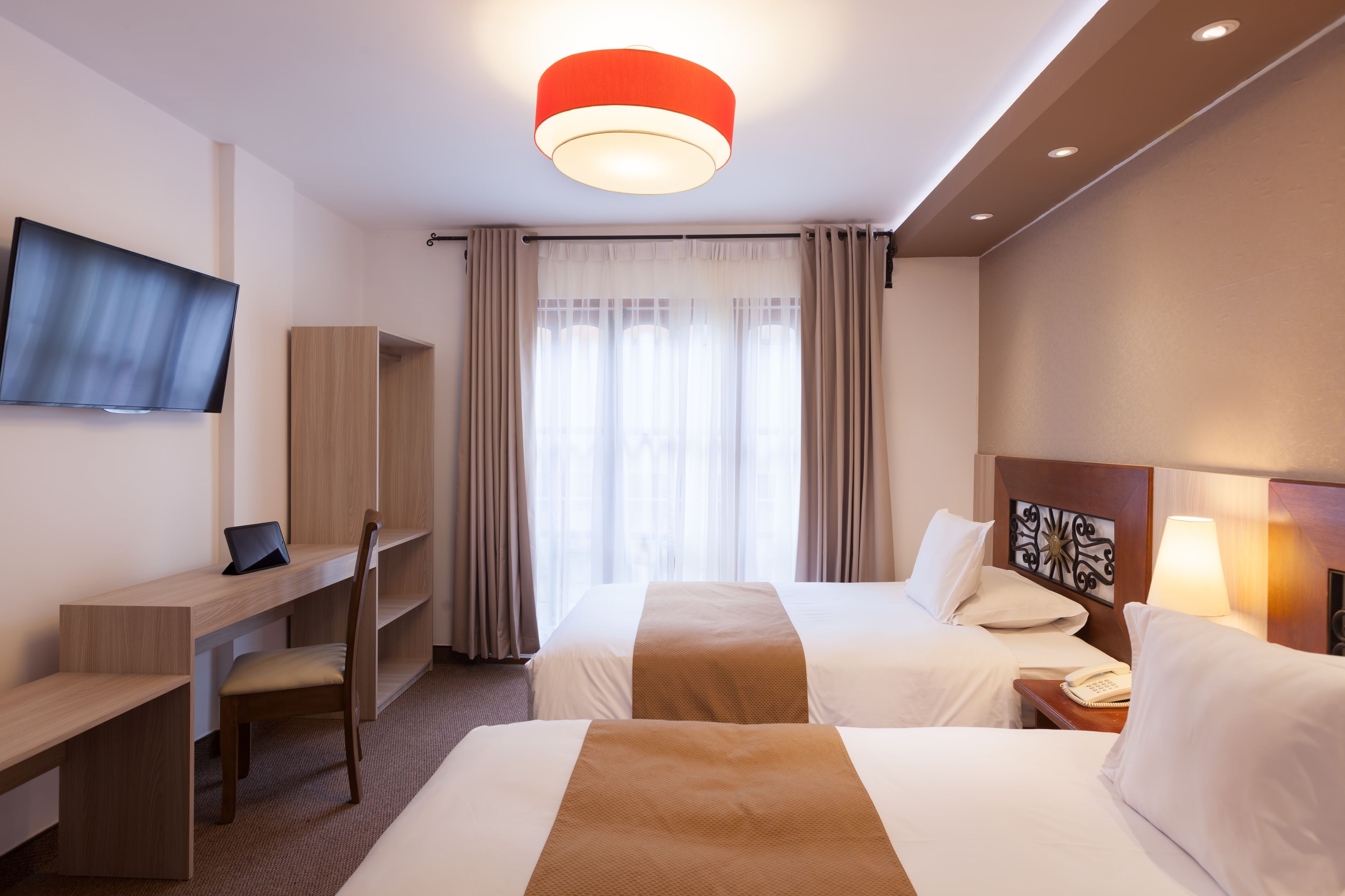 A hotel room with two beds, a desk, a TV, and a window with curtains.