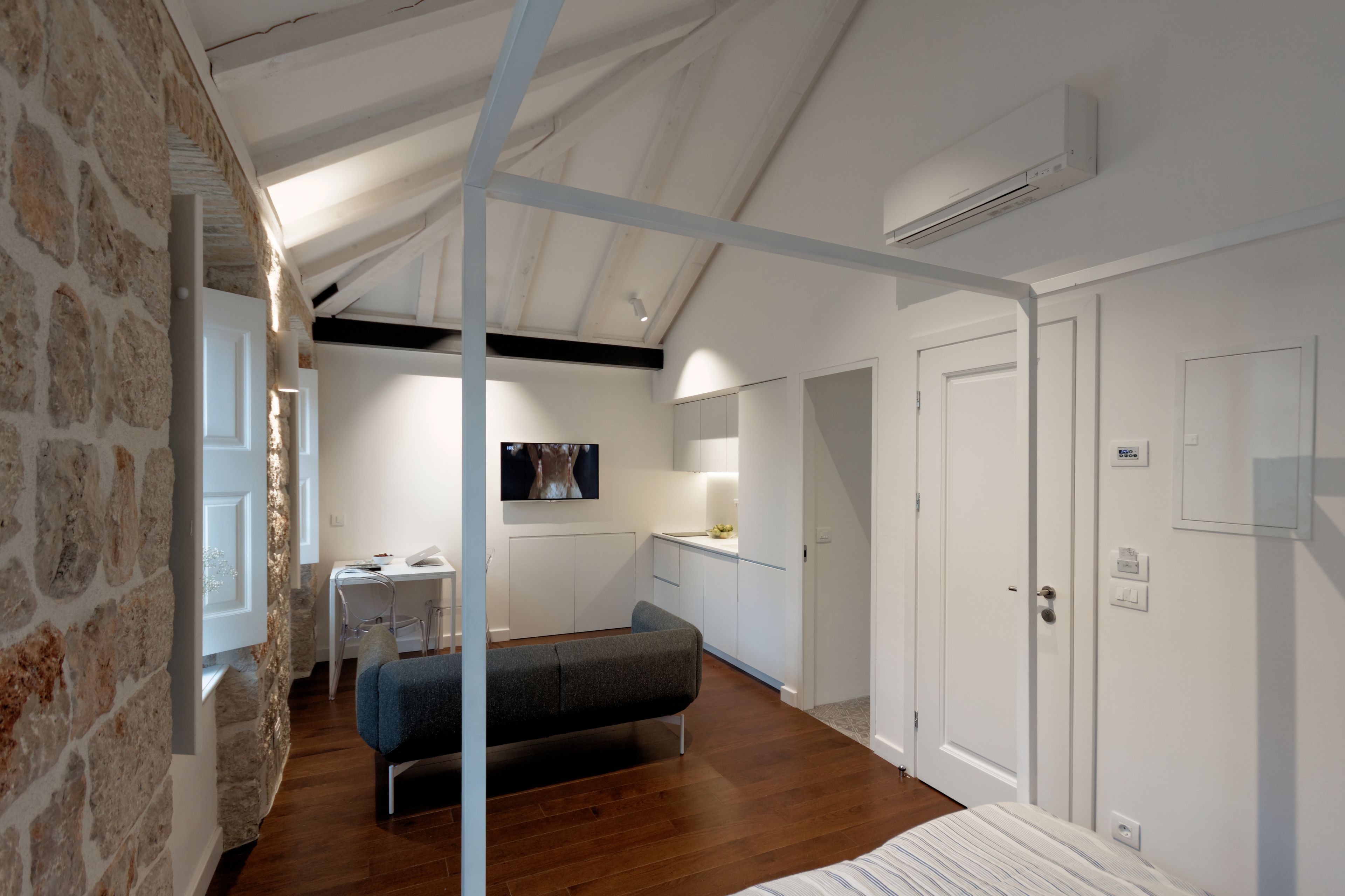 Un dormitorio moderno con pared de piedra, suelo de madera, un banco, cama, televisor y una kitchenette.