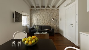 A living room with a stone wall, a dark sofa, a coffee table, and a dining area with a bowl of fruit. - Apartments Eleganca (Dubrovnik)