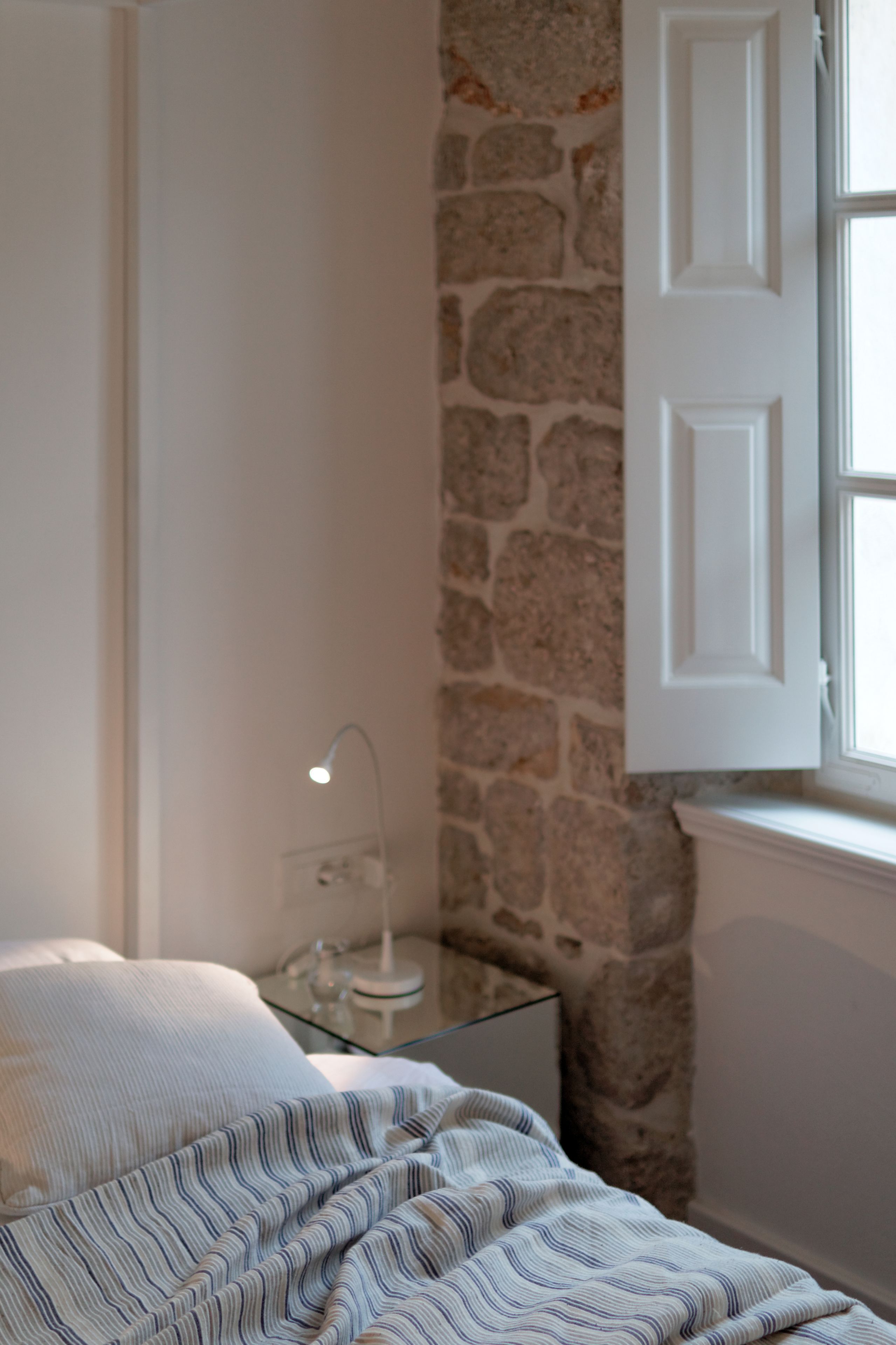 Une chambre à coucher avec un lit, une table de chevet avec une lampe, un mur en pierre et une fenêtre avec une porte blanche.