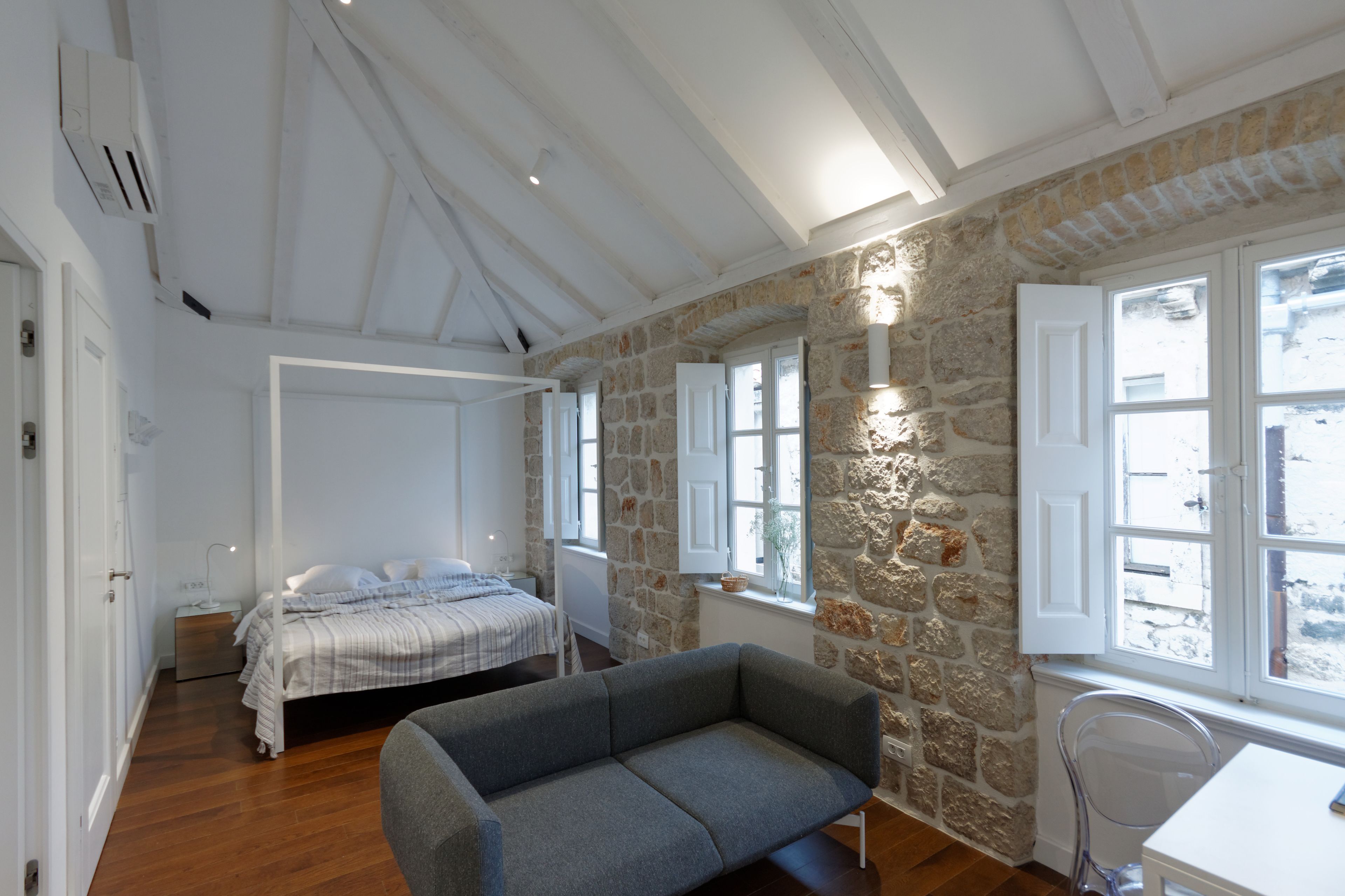 A bedroom with a bed, a grey sofa, stone walls, and large windows.