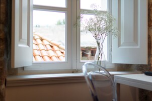 Uma janela com vista para um telhado em cerâmica, um vaso com flores e uma cadeira.
