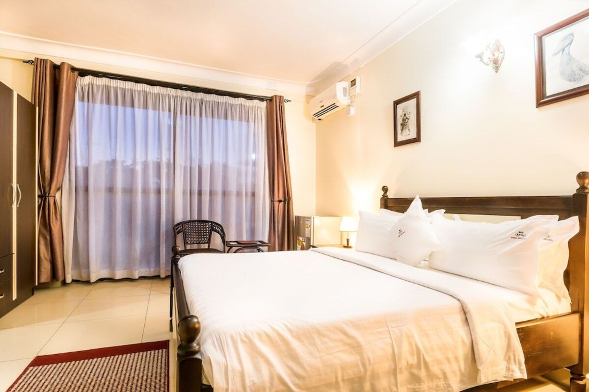 A hotel room with a bed, curtains, a chair, a lamp, and a wall-mounted air conditioner.