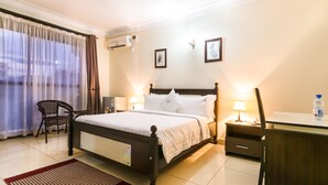 A hotel room with a bed, bedside tables, a desk, a chair, and a window with curtains. - Marie's Royale Hotel (Kampala)