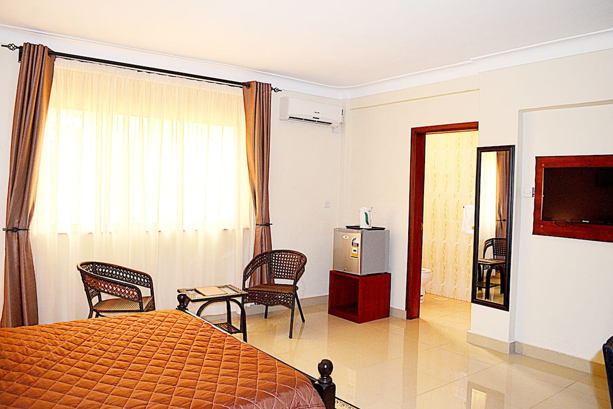 A hotel room with a bed, two chairs, a small table, an air conditioning unit, a television, and a bathroom visible through an open door.