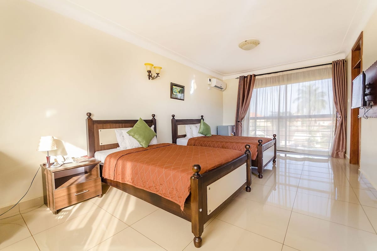 A hotel room with two beds, a bedside table with a lamp, a wall-mounted air conditioner, and a large window with curtains.