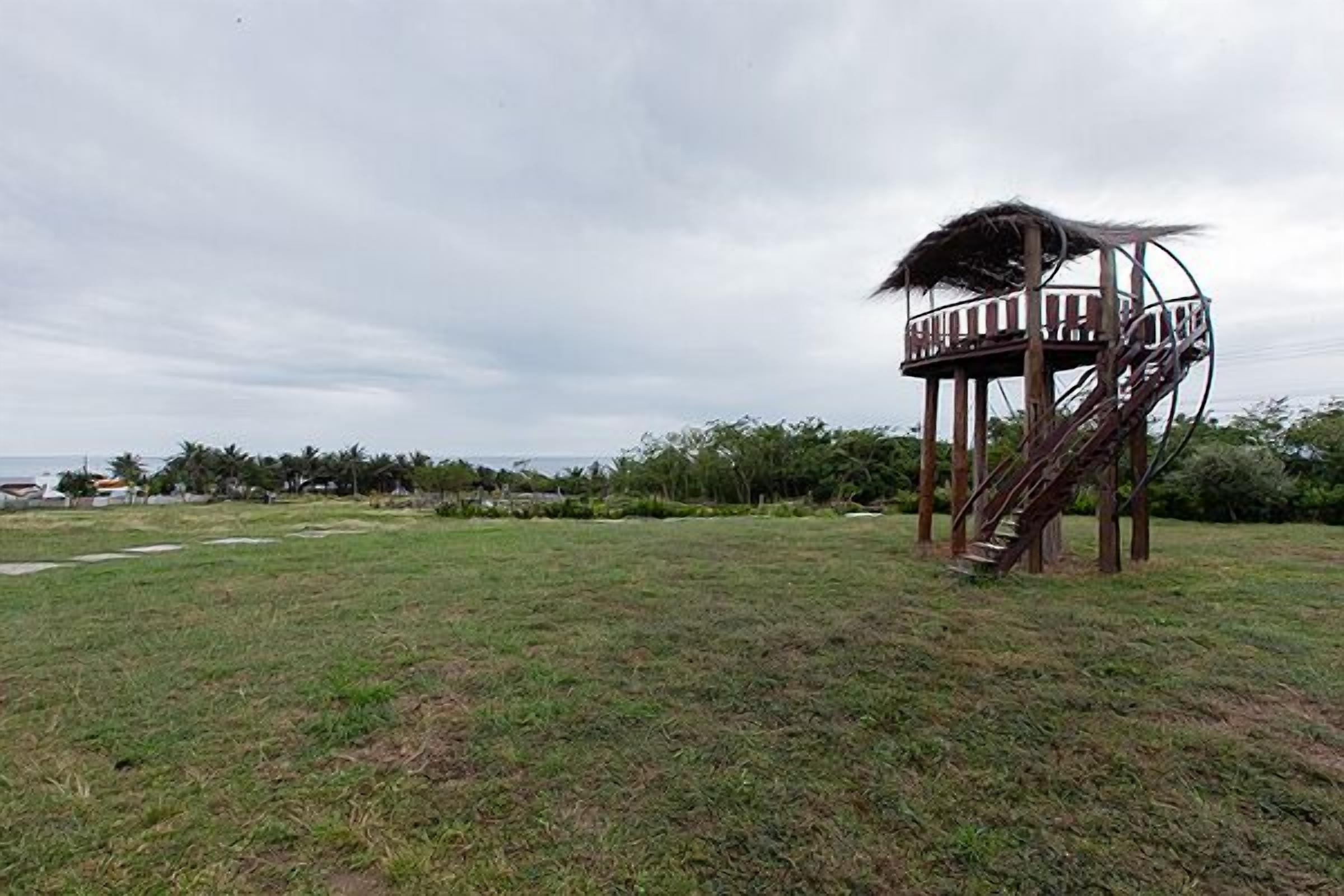 A single-story structure with a spiral staircase and a thatched roof, surrounded by a grassy area and trees.