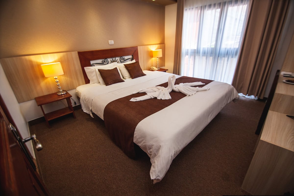 A hotel room with a large bed, two bedside tables, a wooden headboard, and a window with curtains.