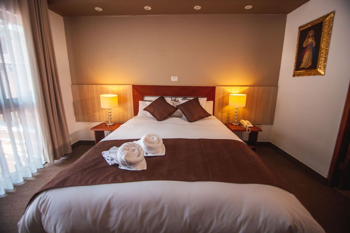 A hotel room with a large bed, two bedside tables with lamps, a painting on the wall, and a window with curtains.