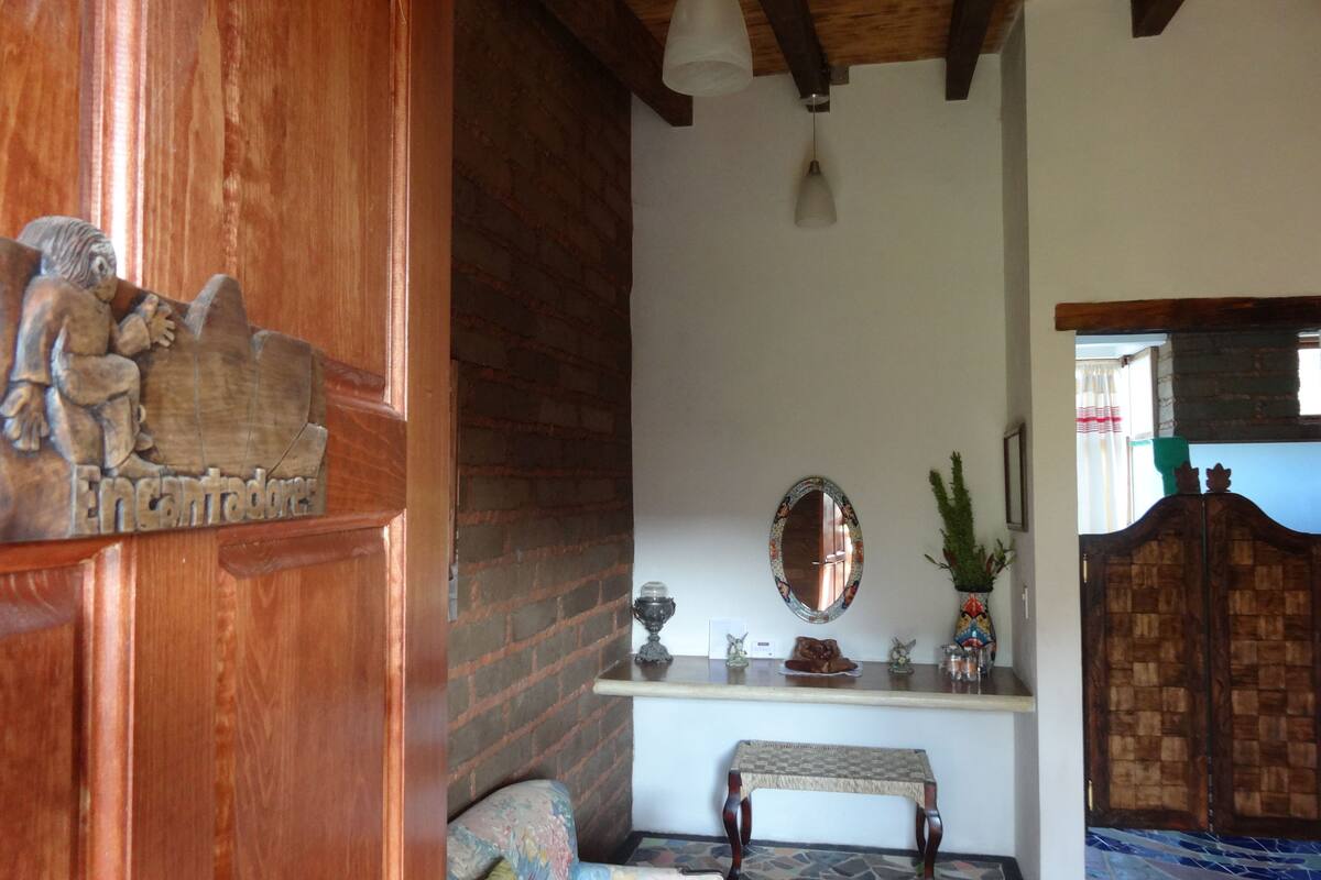 Een houten deur met een houtsnijfiguur, een spiegel, een tafeltje en een bank in een kamer met een betegelde vloer en een houten plafond.