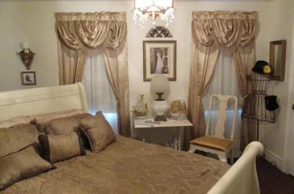 A bedroom with a bed, a chair, a table with a lamp, curtains, a chandelier, and wall-mounted decorations.