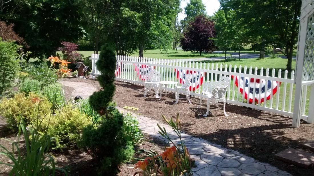 A garden with a white picket fence, a paved pathway, and a small seating area with white chairs and a table.