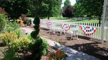 A garden with a white picket fence, a paved pathway, and a small seating area with white chairs and a table.