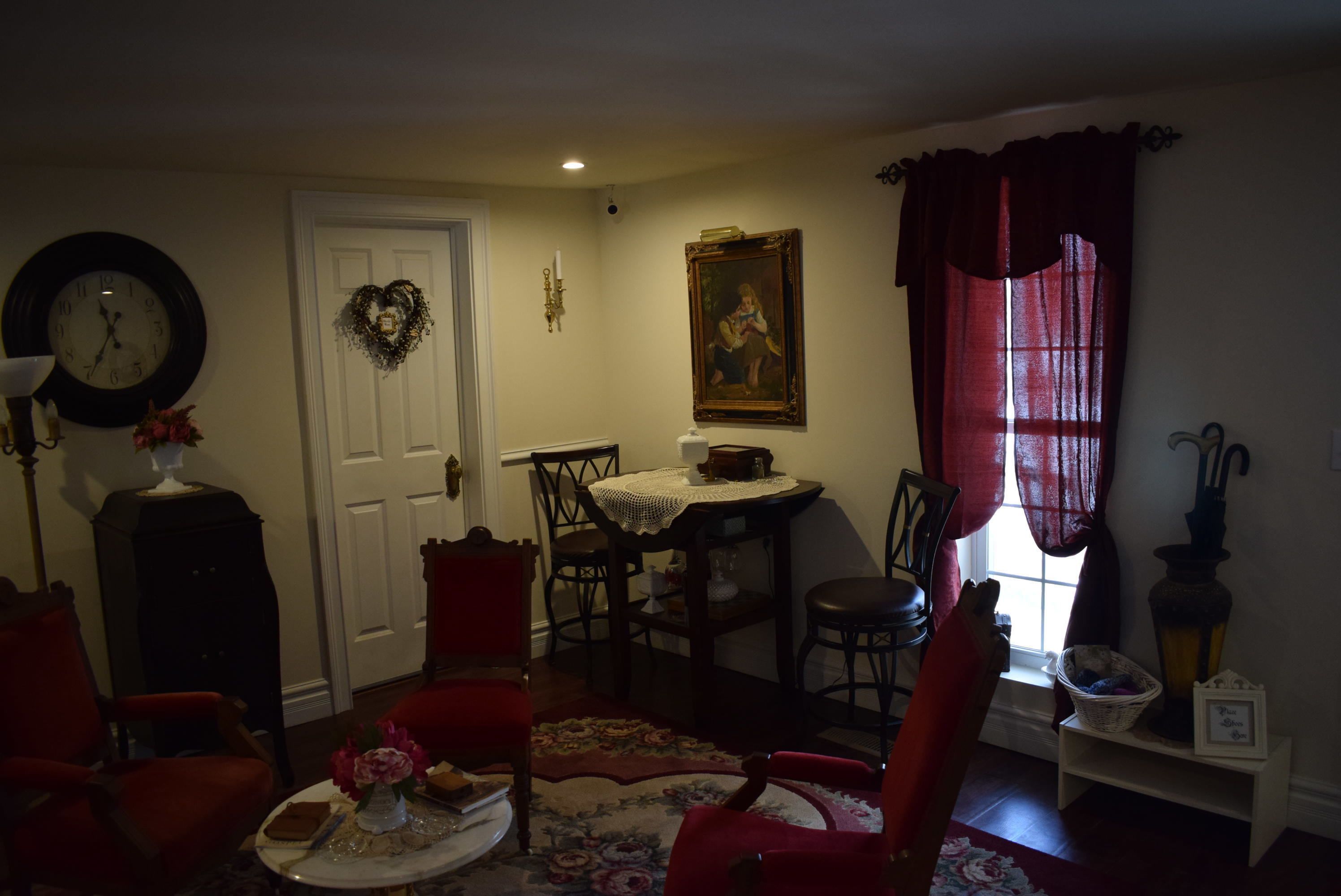A cozy living room with a dining area, featuring a clock, a heart-shaped wreath, a painting, and a window with curtains.