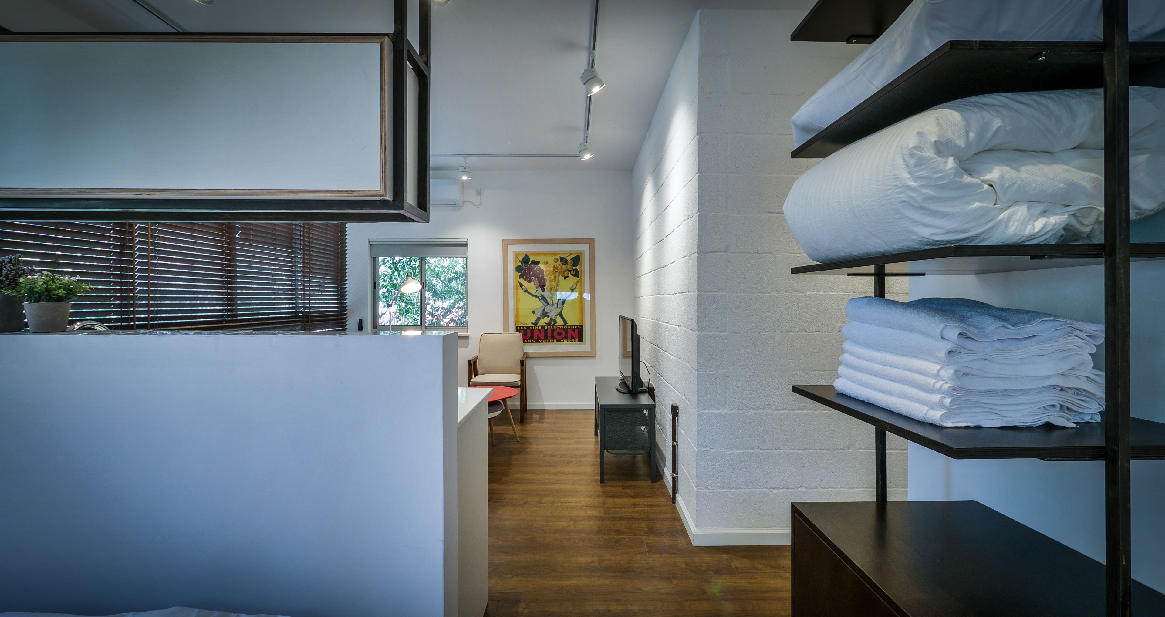 Una habitación de hotel moderna con piso de madera, una cama con ropa blanca, un escritorio con silla y un estante de pared con toallas dobladas.