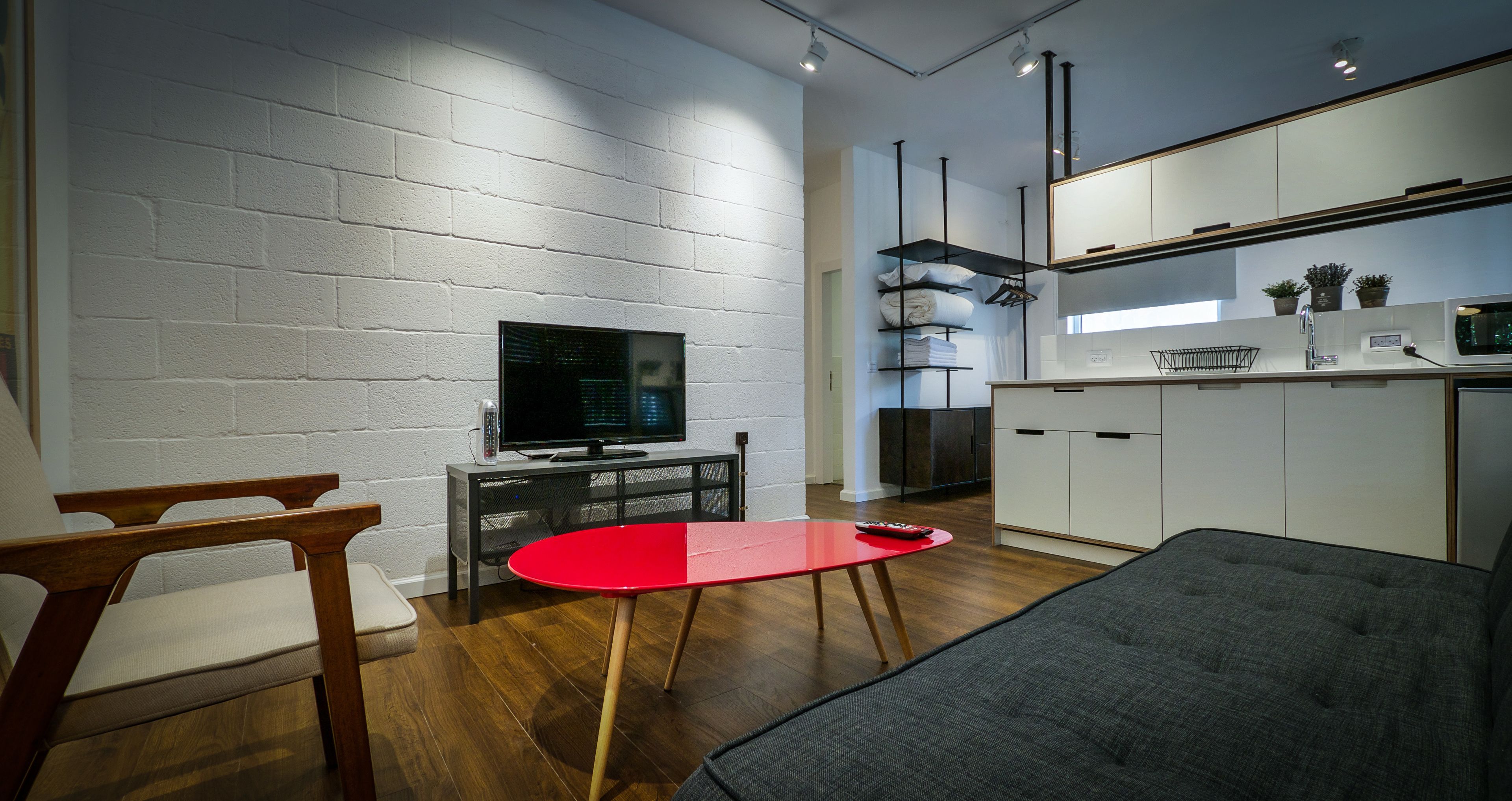 A modern living room with a red table, a grey sofa, and a TV on a stand against a white brick wall.